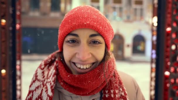 Retrato de mulher alegre feliz em gorro vermelho tricotado e lenço que anda na praça da cidade com iluminações festivas. neve está caindo e flocos de neve voando ao redor. Belo dia de inverno. Neve macia cai — Vídeo de Stock