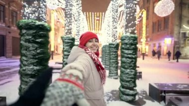 POV first person view of happy woman holding someones hand and leads while walks on city square with festive colorful illuminations. girl rejoices in holiday and decorations. snowflakes flying around