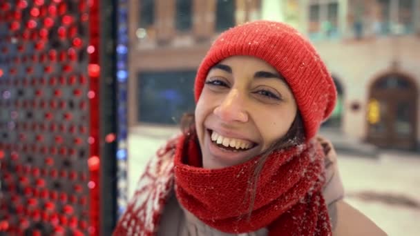 Portrait of happy laughing cheerful young woman in knitted red cap and scarf walking on city square with festive illuminations. snow is falling and snowflakes flying around — Stockvideo
