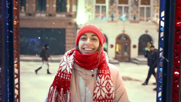 Portrait of happy laughing cheerful young woman in knitted red cap and scarf walking on city square with festive illuminations. snow is falling and snowflakes flying around — Wideo stockowe