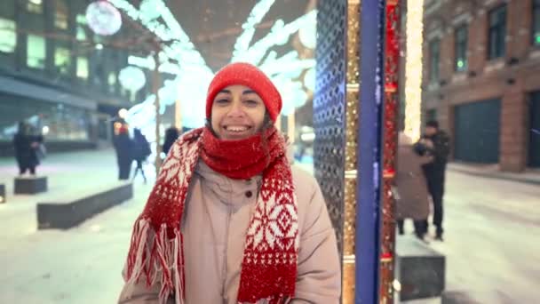 Tiro gimbal de mulher linda sorridente feliz em tricotado boné vermelho e cachecol andando na praça da cidade com iluminações coloridas festivas na noite nova eyar. neve está caindo e flocos de neve voando ao redor — Vídeo de Stock