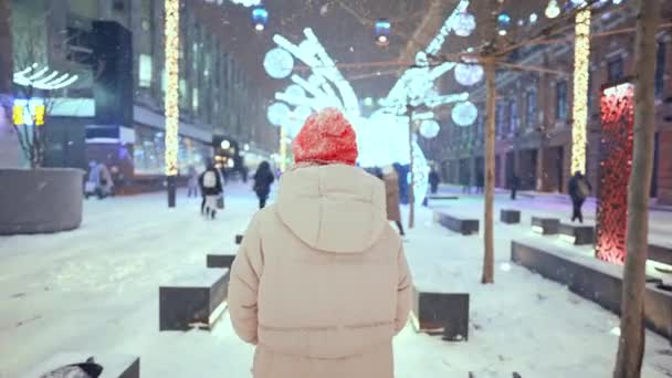 Rear view camera following woman in knitted red cap and scarf walking on city square with festive colorful illuminations at night. soft snow is falling and snowflakes flying around. — Wideo stockowe