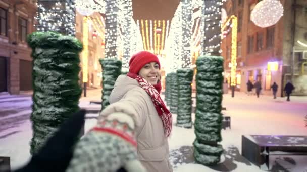 POV primeira pessoa vista de mulher feliz segurando alguém mão e leva enquanto caminha na praça da cidade com iluminações coloridas festivas. menina se alegra em férias e decorações. flocos de neve voando — Vídeo de Stock