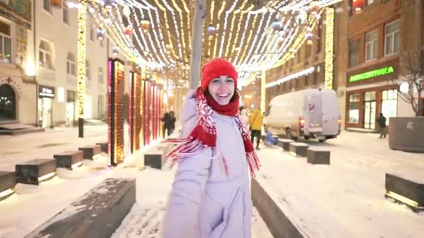 Happy woman in red cap and scarf walks on city square with festive colorful illuminations. girl rejoices in holiday and decorations and whirls. snow is falling and snowflakes flying around — Vídeos de Stock
