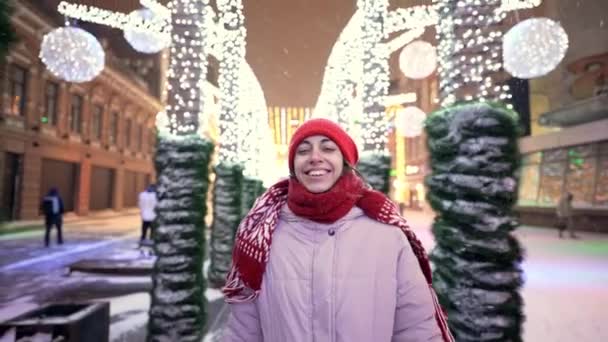 Gimbal Aufnahme von glücklich lächelnden wunderschönen Frau, die auf dem geschmückten Stadtplatz mit festlichen bunten Illuminationen in der neuen Nacht spaziert und die Hände hebt. Schnee fällt und Schneeflocken fliegen herum — Stockvideo