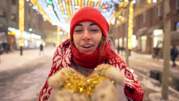 Vidám nő lassított felvételen fúj konfettit a főtéren, ünnepi megvilágítással a téli éjszakákon. Boldog lány, aki elfújja az arany csillámot a kezéről. Esik a hó és hópelyhek röpködnek. — Stock videók