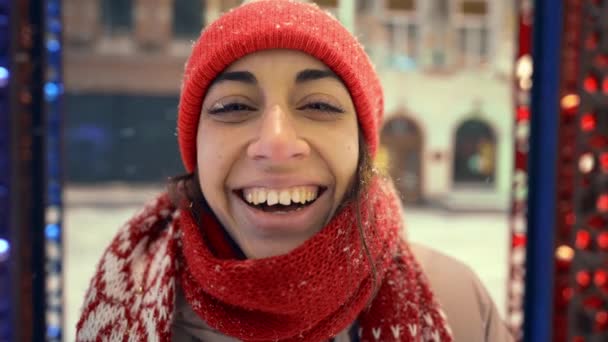 Funny happy laughing cheerful young woman in knitted red beanie and scarf on city square with festive illuminations. snow is falling on cap and eyelashes, snowflakes flying around — Stockvideo