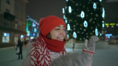 Kırmızı şapkalı ve eşarplı mutlu genç kadının portresi gece şehir meydanında ışıklandırma arka planında halka açık bir Noel ağacına karşı duruyor. Yeni yıl ve kış tatili konsepti
