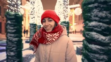 örgü ören kırmızı şapkalı, atkılı, neşeli renkli ışıklarla New eyar gecesinde şehir meydanında yürüyen mutlu, güzel bir kadının görüntüsü. Kar yağıyor ve etrafta kar taneleri uçuşuyor.