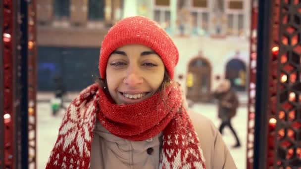 Feliz sorrindo alegre jovem mulher em tricotado boné vermelho e cachecol andando na praça da cidade com iluminações festivas. neve está caindo e flocos de neve voando ao redor — Vídeo de Stock