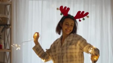 Ren geyiği boynuzu takan genç ve güzel bir kadın. Evde Noel ışıklarıyla dans ediyor.