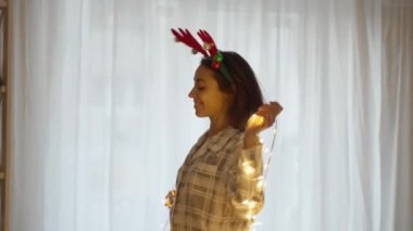 Ren geyiği boynuzu takan genç ve güzel bir kadın. Evde Noel ışıklarıyla dans ediyor.