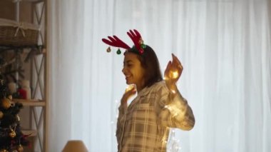 Mutlu, güzel bir kadının portresi pijama giyiyor ve geyik boynuzları takıyor Noel ışıklarıyla Noel ağacıyla evde dans ediyor. xmas ve mutlu yıllar konsepti