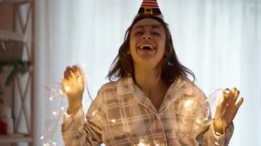 Pijama giyen mutlu ve güzel bir kadının 4 yavaş çekimde portresi ve Noel ağacıyla evde Noel ışıklarıyla dans eden küçük Elf şapkası. xmas ve mutlu yıllar konsepti