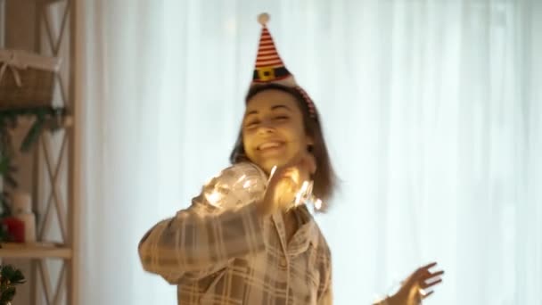 4k slow motion portrait of happy cheerful pretty woman wearing pajama and little Elf Cap headband dancing with Christmas lights at cozy home with christmas tree. xmas and happy new year concept — Stock Video
