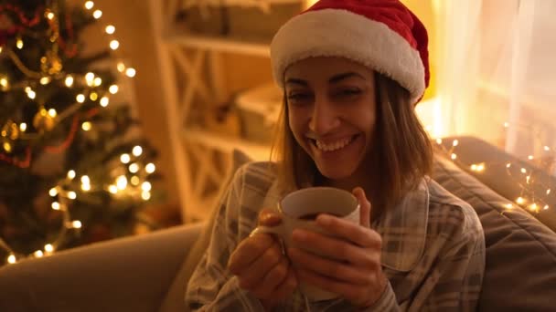 Женщина в пижаме и красной шляпе Санты сидит на диване, смотрит телевизор и пьет горячий шоколад из кружки, расслабляется в канун Рождества, Новый год. девушка в уютном украшенном доме с огнями и рождественской елкой — стоковое видео