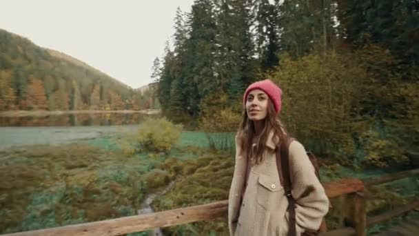 Hübsches Hipster-Mädchen in Freizeitkleidung und Mütze mit Rucksack Wandern im Wald im Herbst. Touristischer Backpacker bei einer Wanderung im Wald am Seeufer. Wanderin spaziert auf Holzbrücke am Bergsee — Stockvideo