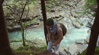 Sırt çantalı bir adam ormanda nehir kenarında geziyor. Yolunu kaybetmiş bir turist. Vahşi doğa güzelliği, sağlıklı aktif yaşam tarzı