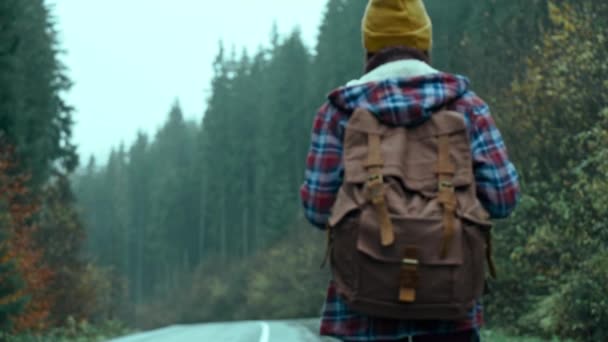 Vista posterior mujer autoestopista viajero con mochila camina camino analógico al aire libre en frío húmedo niebla día de otoño y espera un coche. viaje de viaje enganche montaña naturaleza vacaciones viajes turismo libertad — Vídeo de stock