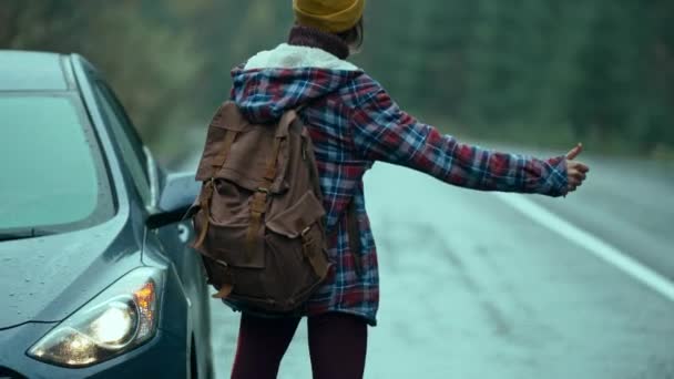 Tylny widok kobieta podróżnik z plecakiem stoi w zepsutym samochodzie na poboczu drogi w zimny mokry mglisty jesienny dzień i próbuje zatrzymać samochód. kobieta kierowca czeka na pomoc na drodze — Wideo stockowe