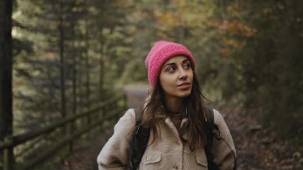 Mulher caminhante sorridente em gorro rosa Caminhadas na floresta no outono. Menina caucasiana saudável ativo com mochila a fazer caminhadas em madeira. viajante feminino caminha na floresta, lazer, bio-turismo — Vídeo de Stock