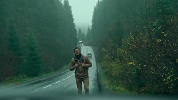 Człowiek autostopowicz podróżnik z plecakiem spacery alog drogi na zewnątrz w mglisty, mokry jesienny dzień i czeka na samochód. mężczyzna rozgląda się dookoła i prosto w aparacie — Wideo stockowe