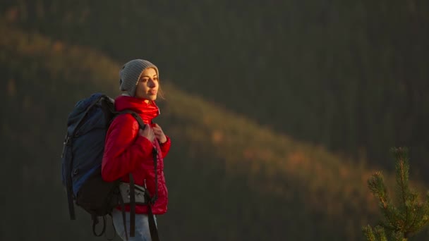 Potret seorang wanita muda yang tersenyum dengan jaket merah dan kupluk terlihat percaya diri dan bahagia dengan ekspedisi hiking-nya di luar ruangan di puncak gunung saat matahari terbenam — Stok Video