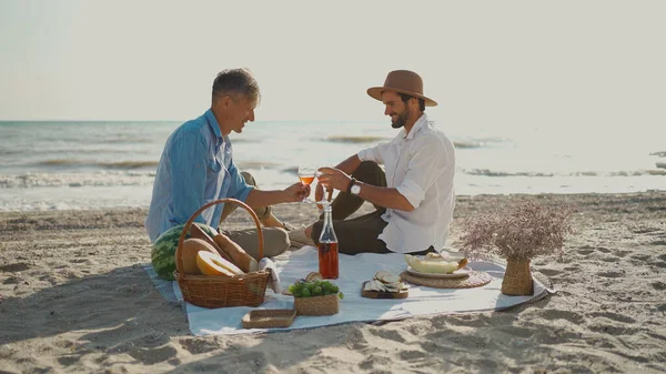 Aikuinen homo pari nauttia romanttinen päivämäärä piknik rannalla, hurraavat lasit viiniä — kuvapankkivalokuva