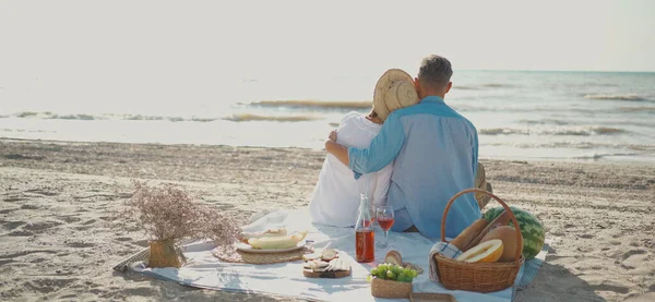 Voksne homoseksuelle par som sitter, omfavner og nyter romantisk piknik på stranden – stockfoto