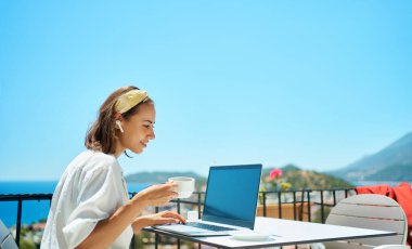 Yetişkin bir kadın dizüstü bilgisayar kullanıyor. Kahve içiyor. Tatil köyünde çalışmaktan keyif alıyor.