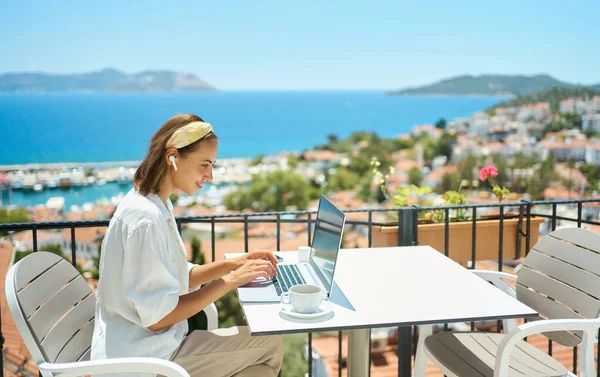 Nuori nainen, joka käyttää kannettavaa tietokonetta kahvilan parvekkeella hotelli merinäköalalla — kuvapankkivalokuva