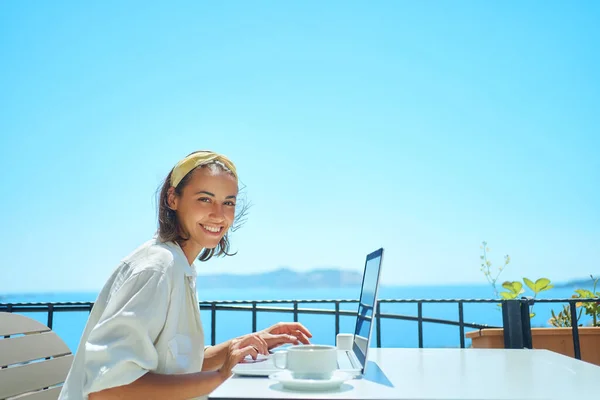 Joven mujer de raza mixta sonriendo a la cámara disfrutando de usar el ordenador portátil en la cafetería resort al aire libre —  Fotos de Stock