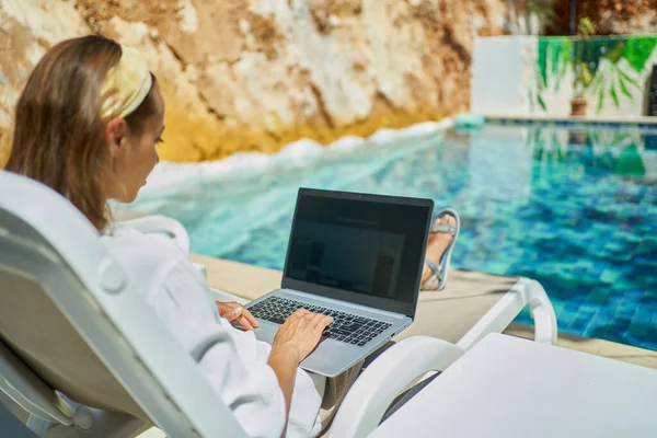Nainen, joka käyttää kannettavaa tietokonetta internetissä, kun makaa aurinkotuoleilla uima-altaalla — kuvapankkivalokuva