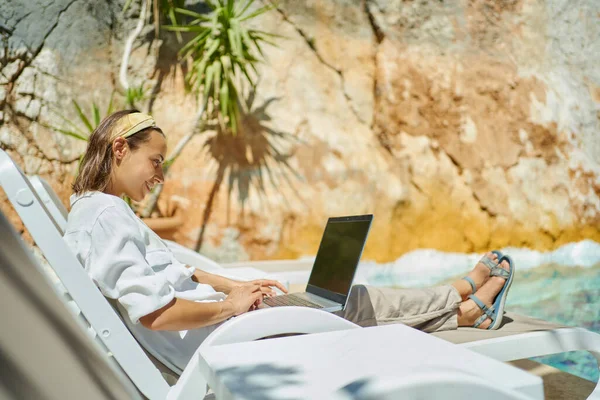 Счастливая женщина фрилансер, использующая ноутбук, печатающая электронные письма, пока лежит у бассейна — стоковое фото