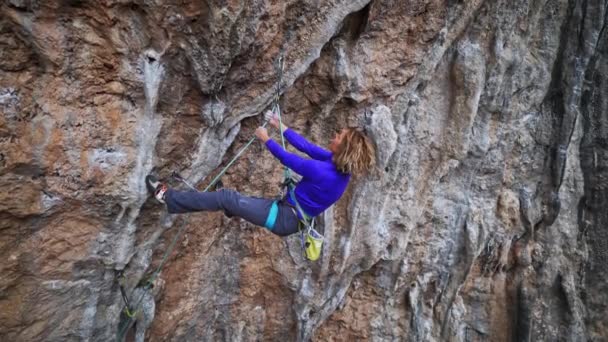 Slow motion smiling tired woman rock climber hanging on rope on hard challenge route on overhanging cliff, resting and chalking hands — Stock Video