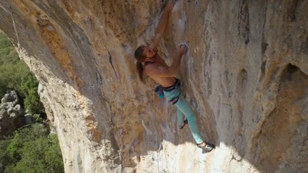 長い髪を持つ若い強力なアスレチックマンロッククライマーは、長い崖の上に登る。男は長いハード移動を作る非常に難しいルートを登る。極端なスポーツ岩登り屋外で — ストック動画
