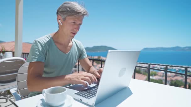 Retrato adulto hombre de pelo gris usando computadora portátil escribiendo correos electrónicos enviando mensajes disfrutando trabajando desde la cafetería del resort con hermosa vista al mar. — Vídeo de stock