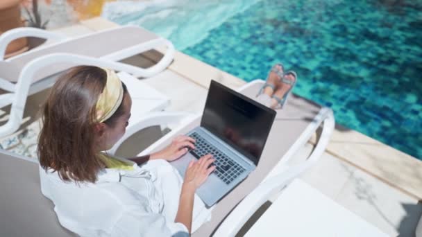 Gesichtslose Frau nutzt Laptop-Computer im Internet, während sie auf Liegestühlen am Pool des Resorthotels liegt — Stockvideo