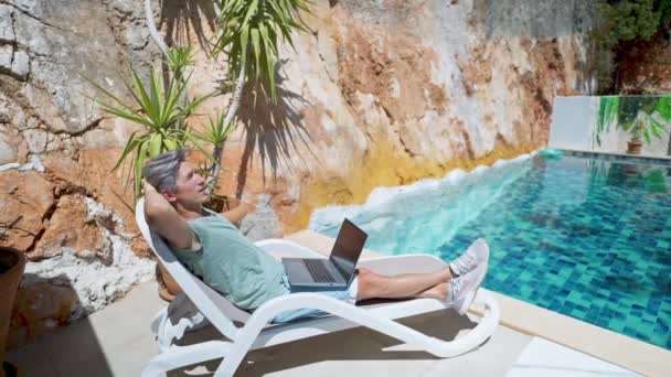 Side view relaxing adult man chilling by the pool during vacation at resort hotel, watching news or video on laptop empty screen — Stock Video