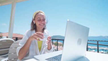 Türk tatil beldesindeki deniz manzaralı terasta oturup sabahları kahve içen memnun bir kadın, bilgisayarını uzaktan çalışma ve serbest konsept olarak kullanıyor.