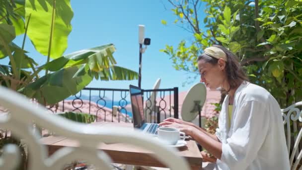 Вид збоку молода жінка-підприємець використовує ноутбук на відкритому повітрі на терасі з пальмами, віддалений онлайн-працює під час літніх канікул — стокове відео