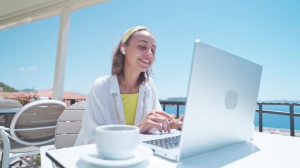 Счастливая молодая женщина носит наушники, общаясь по конференц-связи, сидя на летней террасе с прекрасным видом, видеочатом на собеседовании или дистанционном языковом курсе.. — стоковое видео