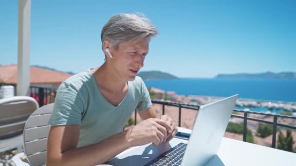 Specialista uomo di mezza età in auricolari wireless con videochiamata sul computer portatile, teleconferenza di lavoro. Millennial man manager o freelancer che lavorano durante le vacanze in località di mare — Video Stock