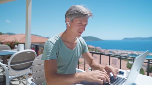 Specialista uomo di mezza età in auricolari wireless con videochiamata sul computer portatile, teleconferenza di lavoro. Millennial man manager o freelancer che lavorano durante le vacanze in località di mare — Video Stock