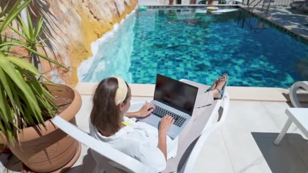 Cámara moviéndose a una mujer disfrutando de relajarse junto a la piscina con ordenador portátil con pantalla blanca y vacía de navegación en línea durante las vacaciones en el hotel resort — Vídeos de Stock