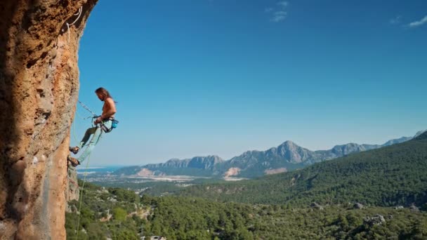 Side view of strong attractive man rock climber getting ready to descend from vertical cliff on top rope. rocky mountain ridge and clear blue sky on background. healthy lifestyle and sport activity — Stock Video