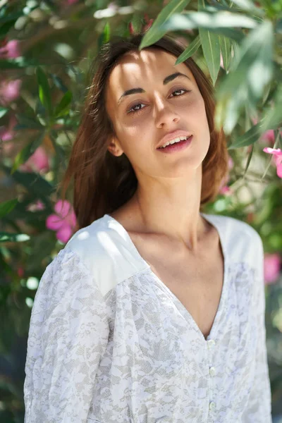 Vakker ung kvinne med perfekt hud, kledd i hvit kjole, som poserer nær rosa blomster. – stockfoto