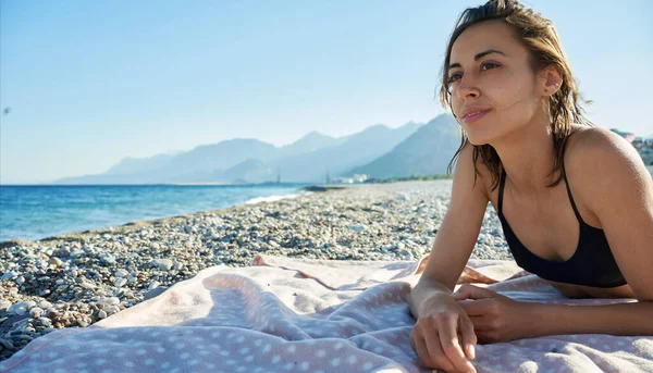 Красивая вдумчивая женщина лежит на морском пляже с горами и смотрит в сторону — стоковое фото