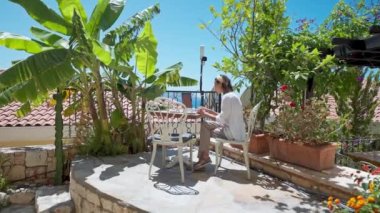 Yan görüş genç bayan iş kadını dışarıda, terasta, palmiye ağaçlarıyla birlikte dizüstü bilgisayar kullanıyor, yaz tatilinde uzaktan kumandalı çalışıyor.