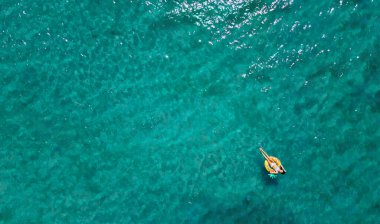 Turkuaz deniz suyunun havadan görünüşü ve sarı yüzme halkasının üzerinde yüzen kadın.